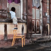 Petit garçon enfant en train de monter sur une chaise de marque DISSI (bois massif de Guyane) et regarde par la fenêtre d'une maison créole.