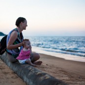 Guyane. Sur la plage des salines (Remire Montjoly) une femme et son bebe en train de regarder le coucher de soleil. Assis sur un tronc de palmier.