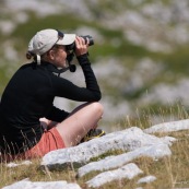 Randonneuse (randonneur) en montagne, femme observant la faune locale avec des jumelles, de trois quart dos, assise sur le sol en montagne. Recherche de chamois ou autres animaux. Observation en milieu naturel. 

Parc National des Abruzzes (Parco Nationale díAbruzzo, Lazio e Molise), Italie.